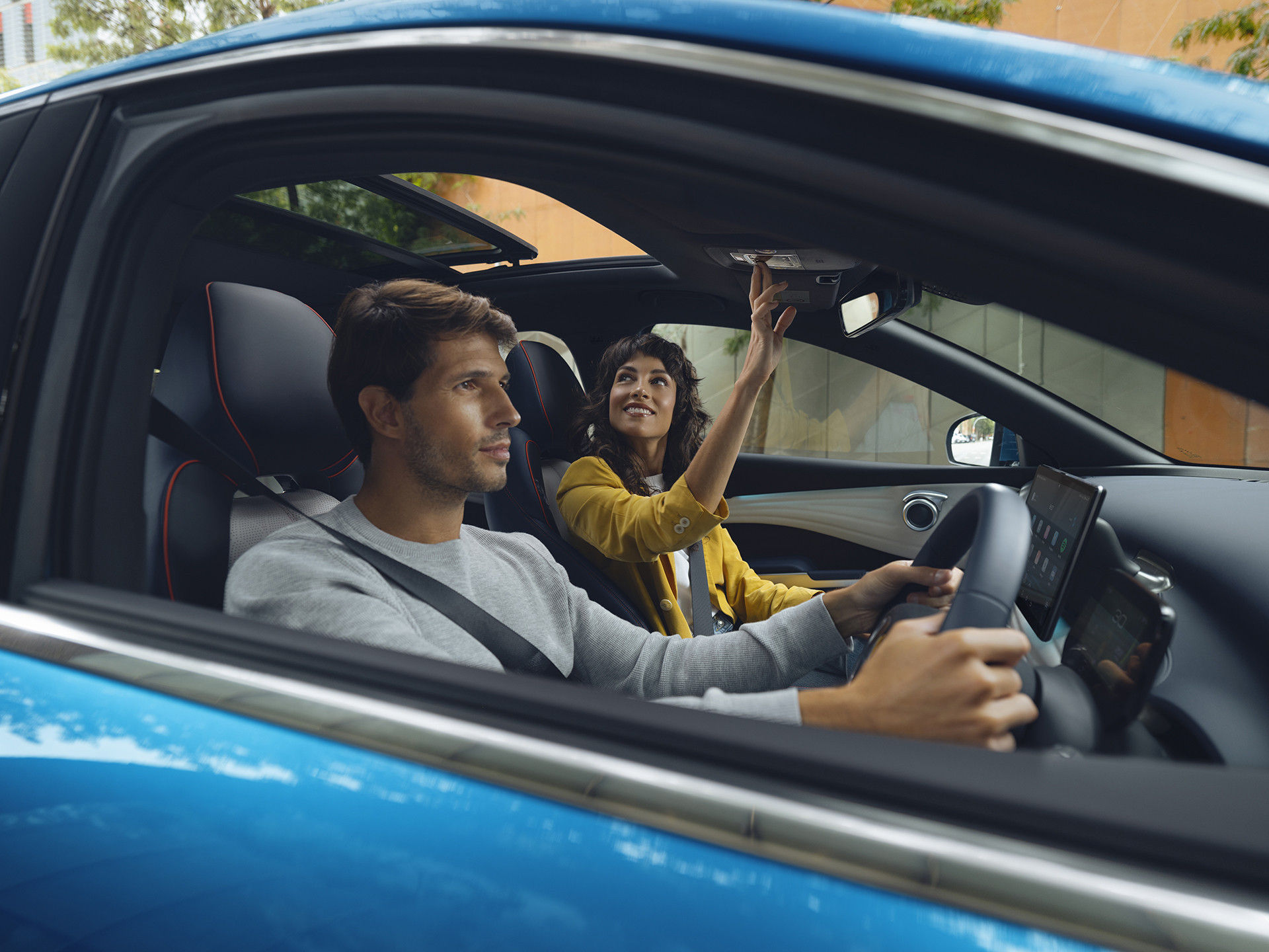 A photograph of a Surfing Blue BYD Atto 3 shot through the drivers window. A man is driving and focusing on the road ahead whilst a passenger (a woman) is operating the electric sunroof