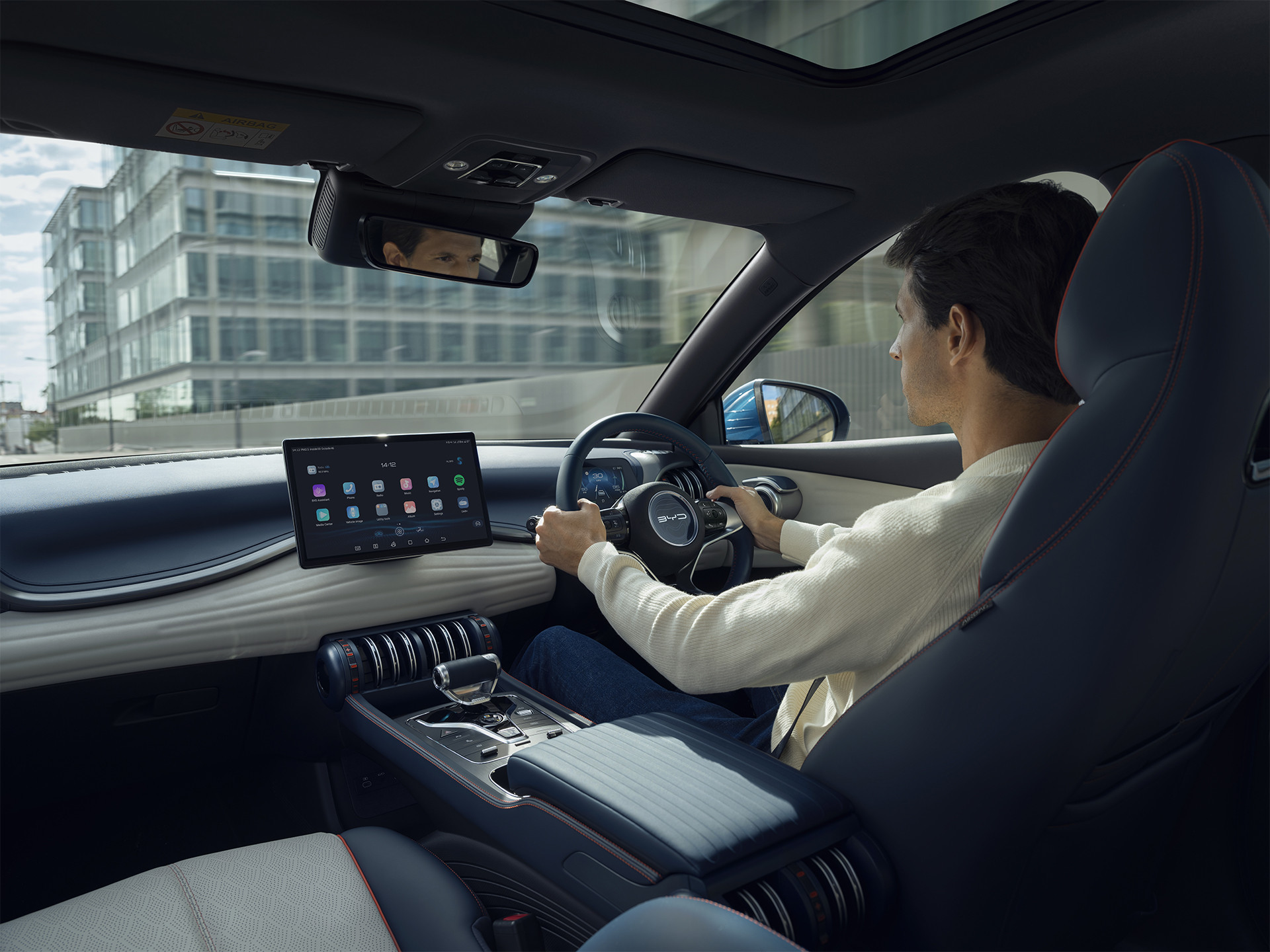 A photograph of the interior of a BYD Atto 3. A man is driving the car. The photo is taken from the rear seats from the perspective of a passenger.